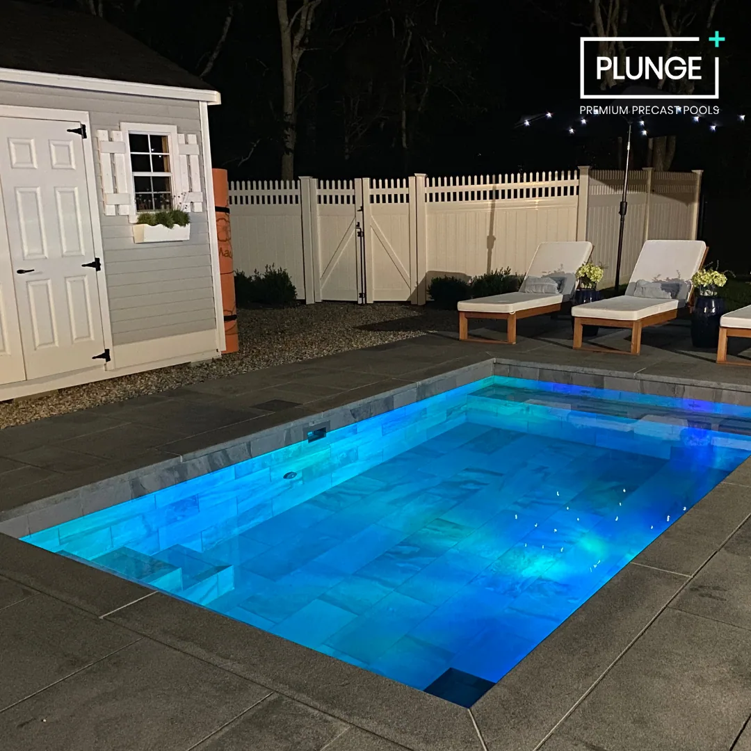 Night view of a backyard swimming pool illuminated with blue lights, surrounded by a concrete deck with lounge chairs and a small shed, enclosed by a white picket fence.
