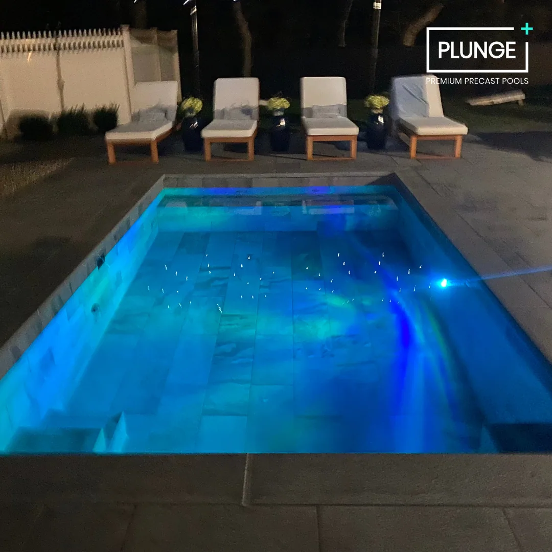 Nighttime view of a small illuminated swimming pool with blue LED lights, surrounded by four lounge chairs with cushions and potted plants, and a privacy fence in the background.