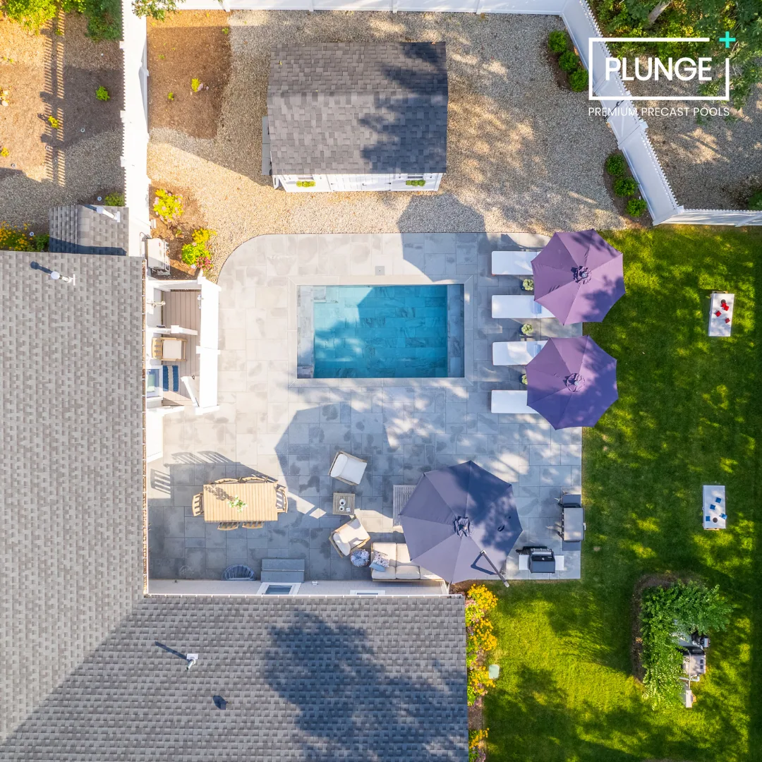 A backyard pool area with four purple umbrellas, lounge chairs, a dining table, and a grassy area with trees and shrubs, surrounded by fences and neighboring houses.