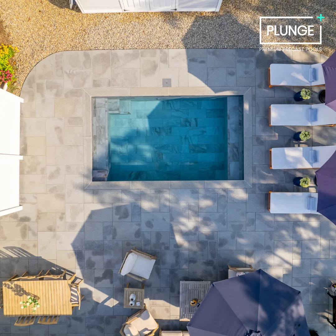 A top-down view of a backyard pool area with four lounge chairs, umbrellas, and small decorative plants, surrounded by tiled paving.