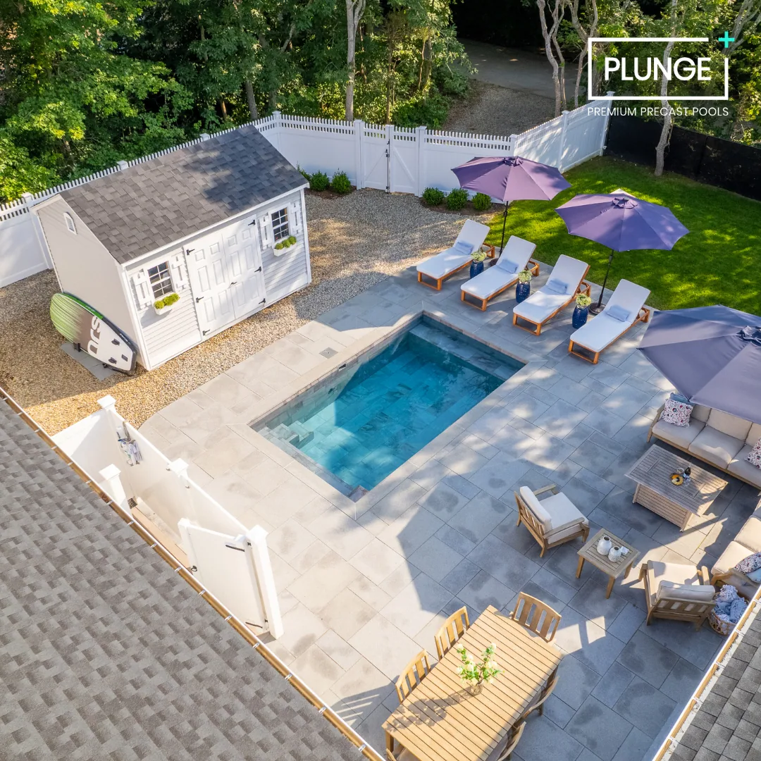 A backyard with a small swimming pool, four white lounge chairs with purple umbrellas, a sitting area with beige furniture and pillows, a wooden dining table with chairs, and a white shed in the corner surrounded by a white fence.
