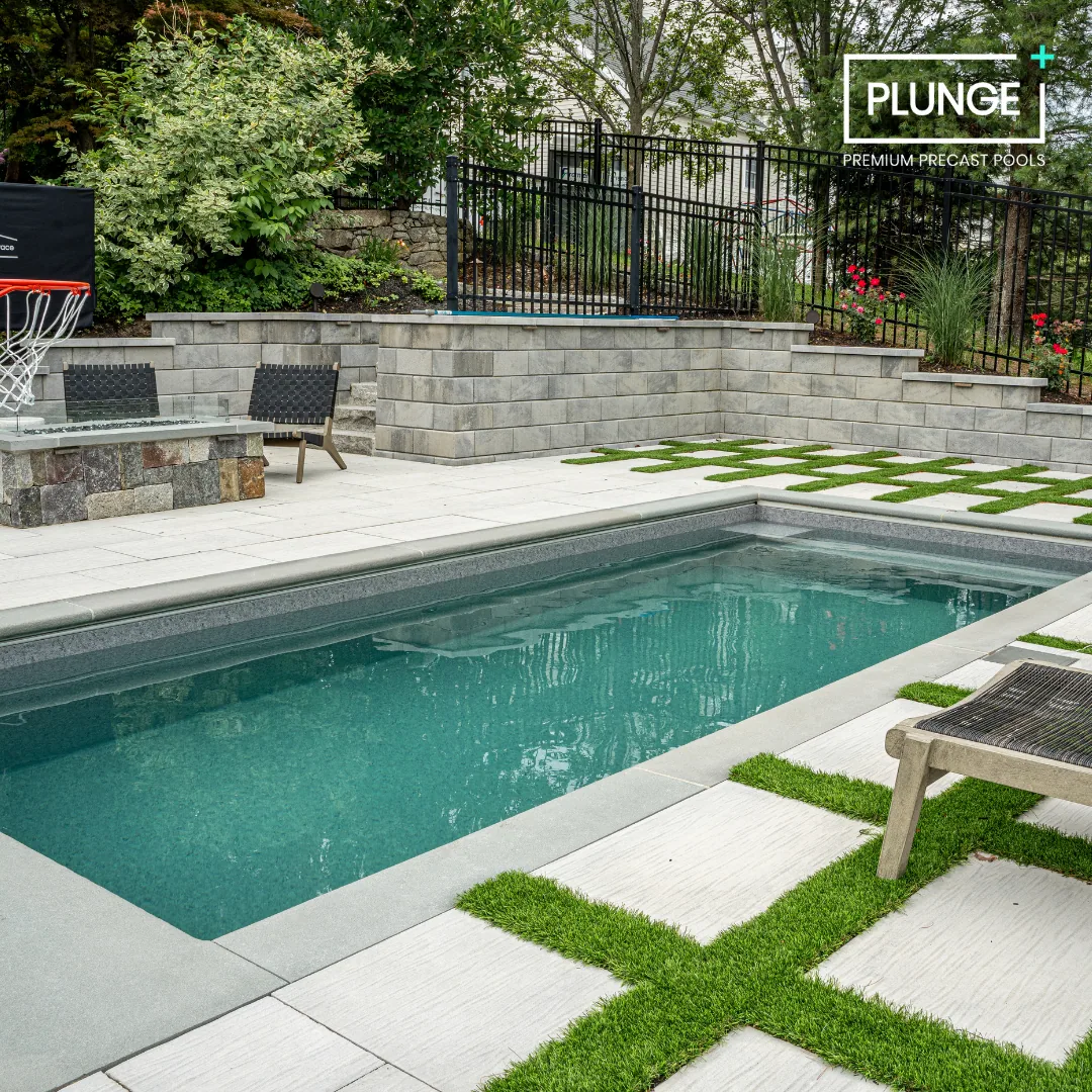A backyard swimming pool with a concrete deck, green grass inlaid in tiles, and a black metal fence around the yard, with trees and bushes in the background.
