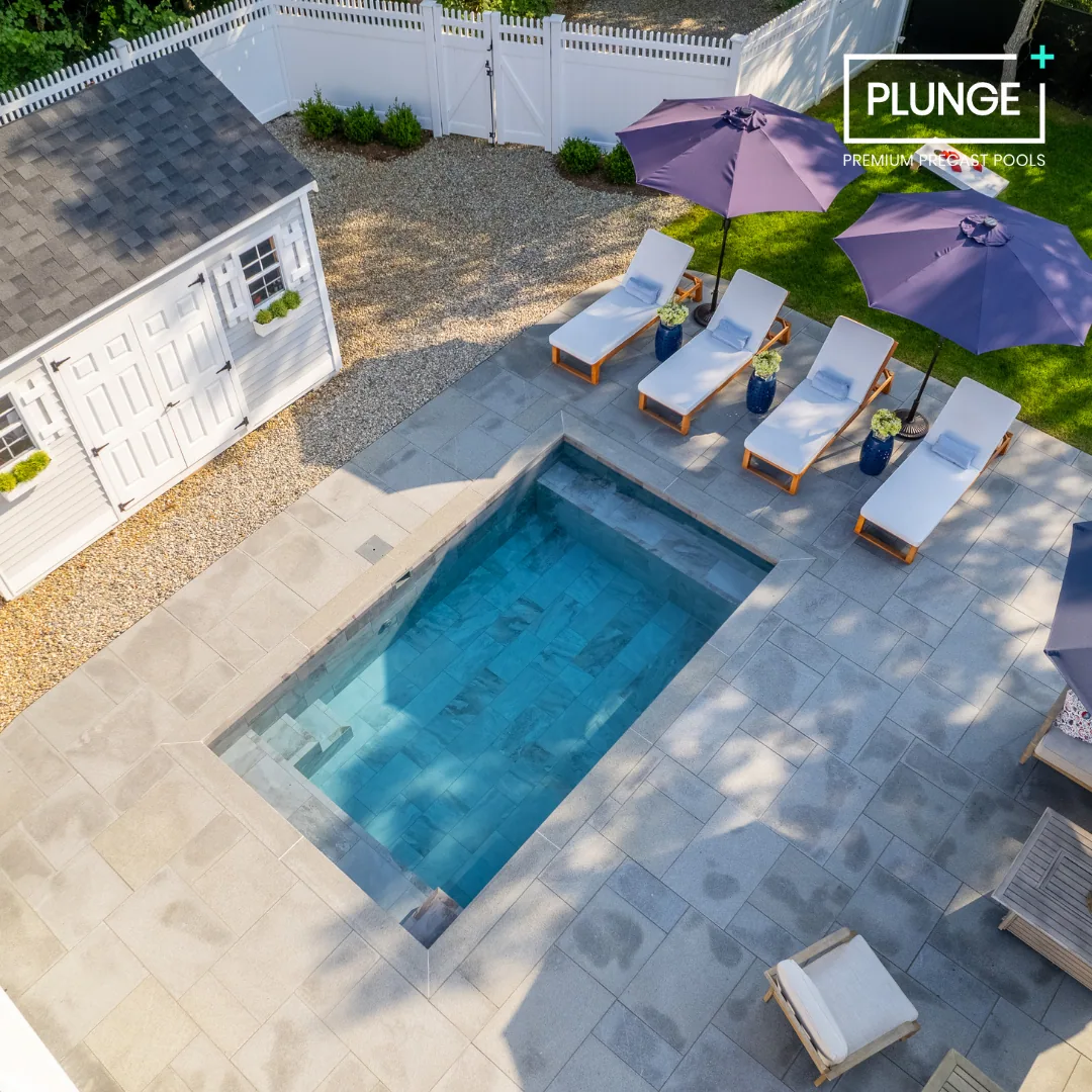 An aerial view of a backyard pool area with four lounge chairs and three purple umbrellas, surrounded by a paved patio, a small shed, and a grassy lawn.