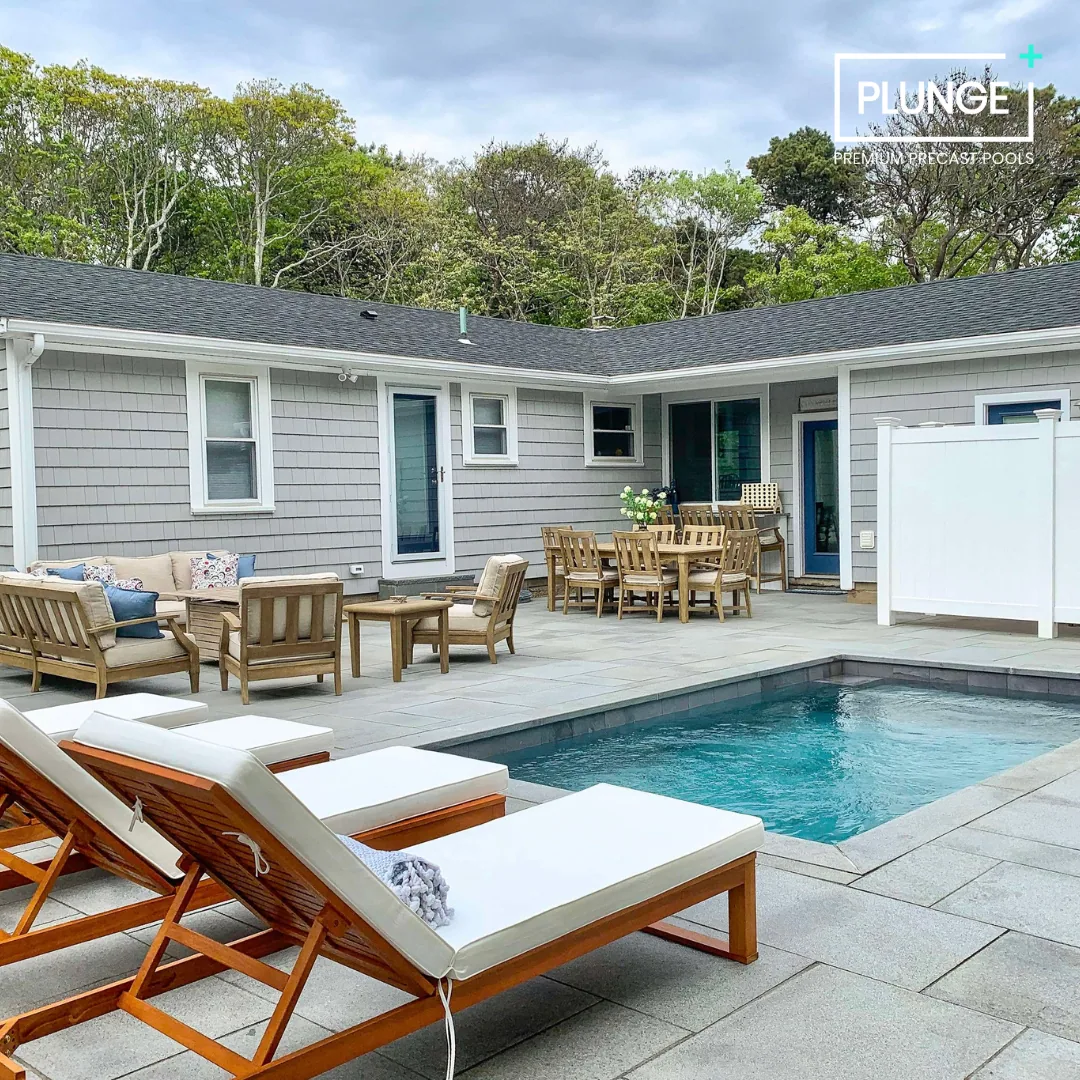 Residential backyard with a small swimming pool, wooden lounge chairs with cushions, outdoor dining table with multiple chairs, surrounding trees, and a house with gray siding and white trim.