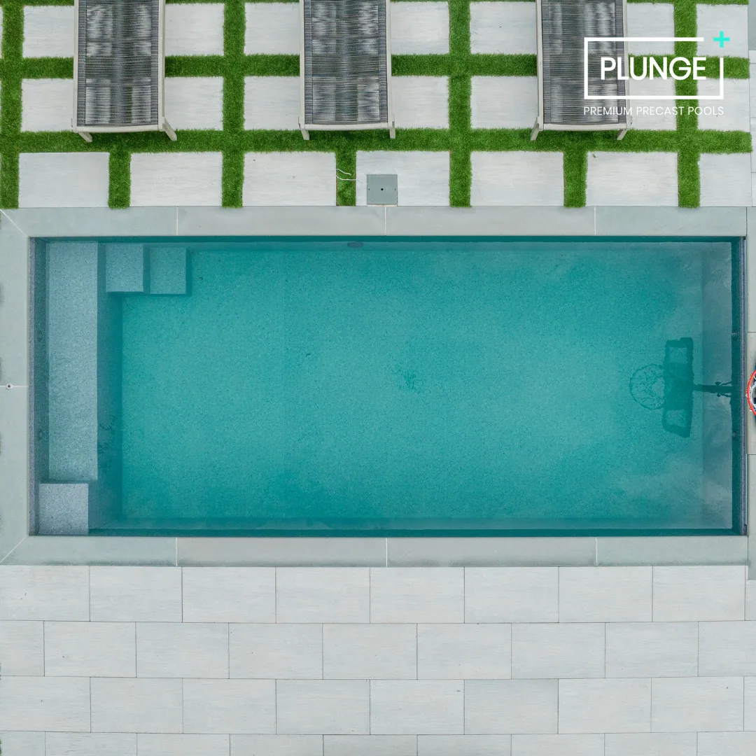 Aerial view of a rectangular in-ground swimming pool with a basketball hoop at one end, surrounded by white tiles and a patio with grass and deck chairs.