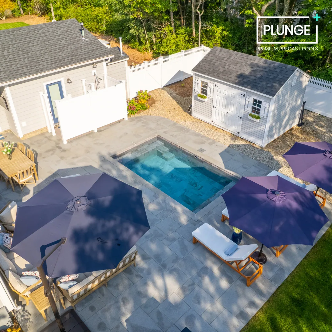 A backyard with a small rectangular swimming pool, outdoor furniture with purple umbrellas, a white shed, a house, and a white fence, surrounded by trees.