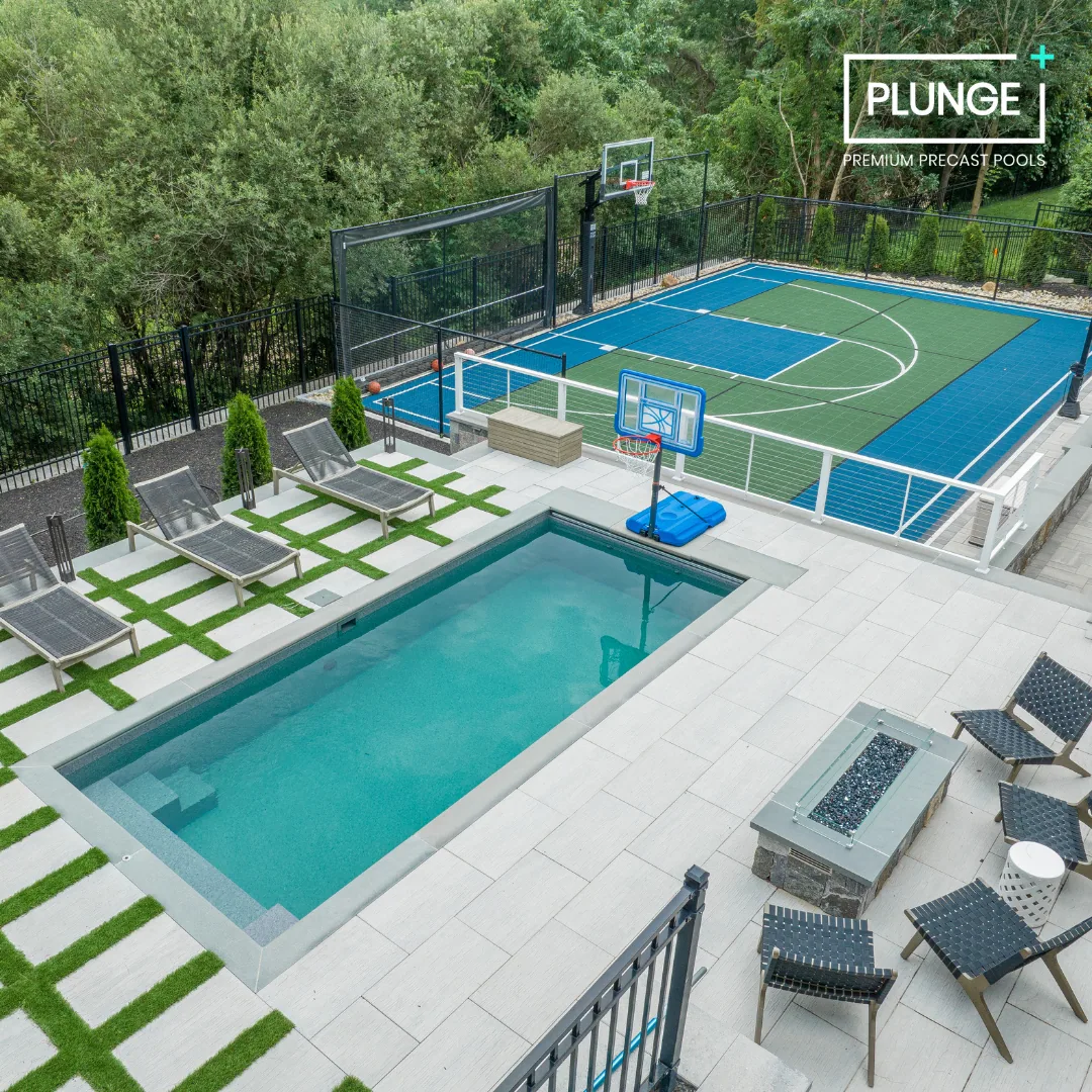 An outdoor backyard featuring a small rectangular swimming pool, a lounge area with six black chairs, a fire pit, a tennis or basketball court with two hoops, and a surrounding green lawn and trees.