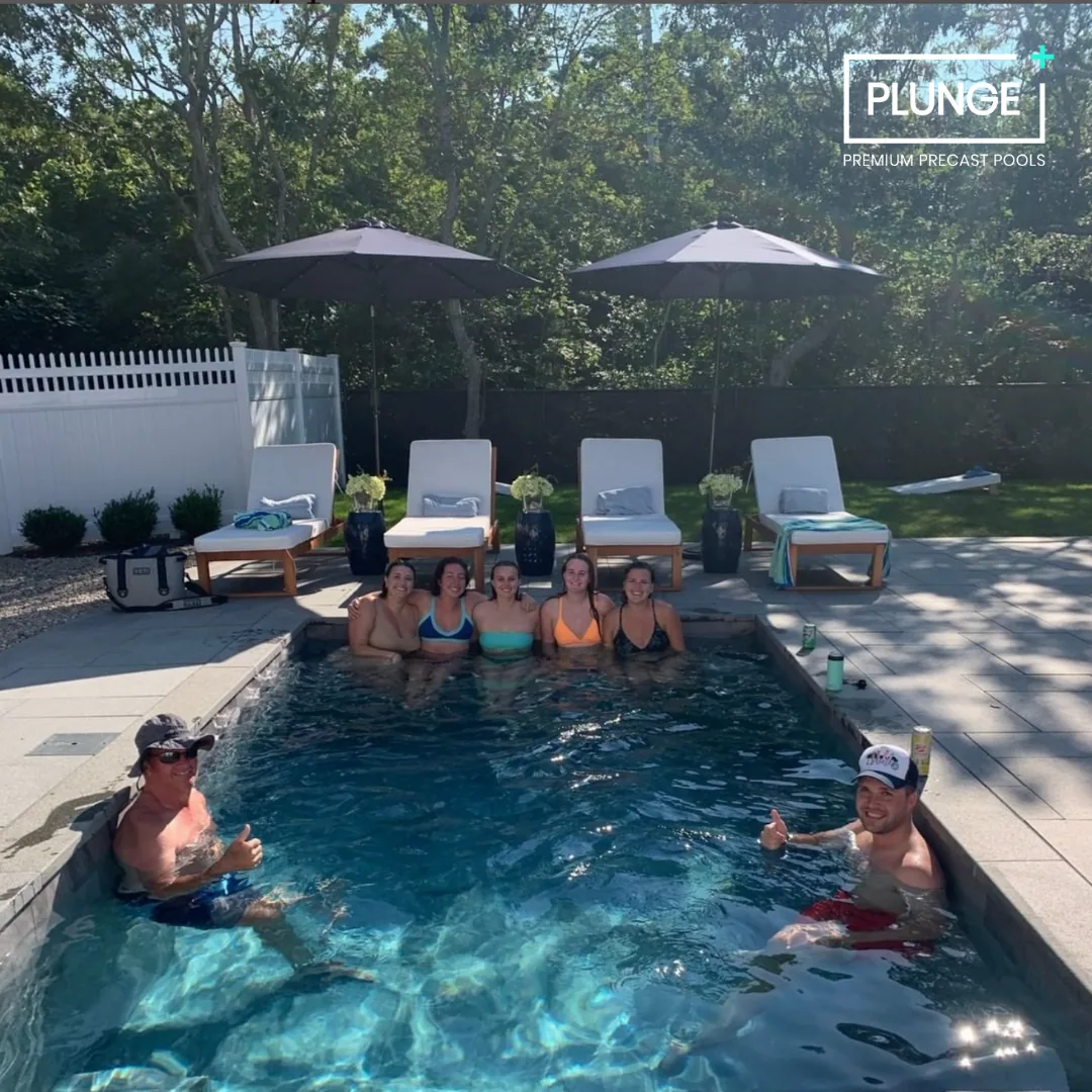 Group of six women and two men enjoying a swimming pool on a sunny day. The women are sitting on the edge of the pool, and the men are in the water giving a thumbs up. The pool area has lounge chairs with umbrellas and a white fence.