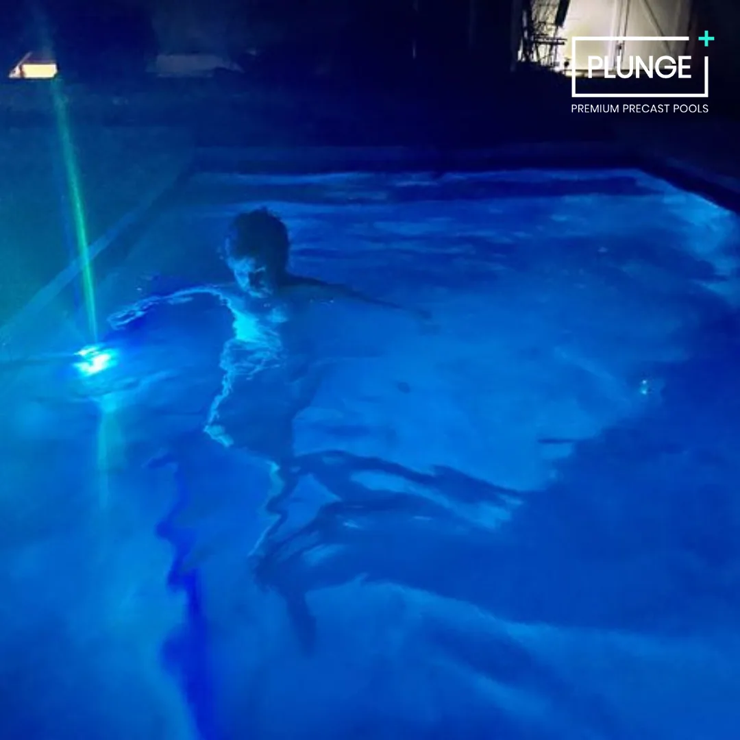 Child swimming in a blue-lit pool at night with a sign that says "PLUNGE PREMIUM PRECAST POOLS."