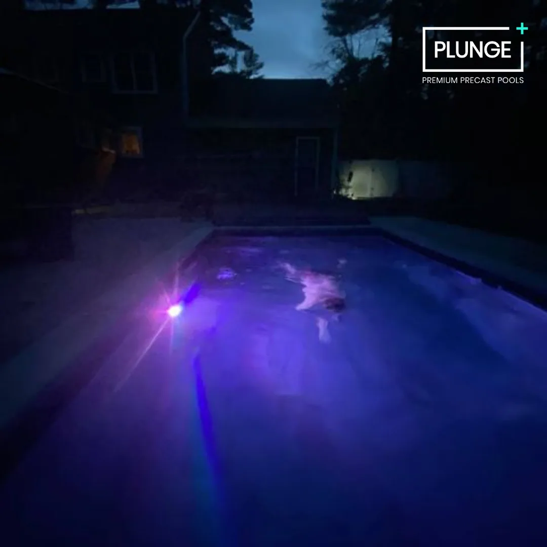 Nighttime view of a swimming pool illuminated with purple and blue lights, with a house and trees in the background.