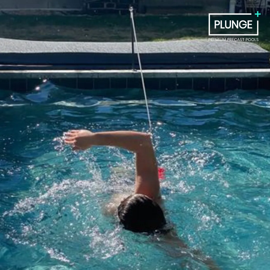 Person swimming in an indoor pool with a black tiled wall and a label that reads 'PLUNGE Premium Precast Pools' in the upper right corner.