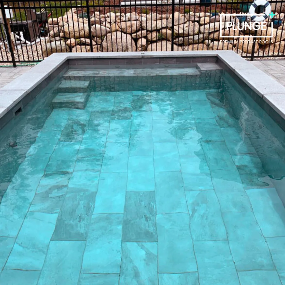 A rectangular private pool with clear water, light blue tiles, built-in steps at one end, and a stone border. A black metal fence surrounds the yard, with rocks and a rocky garden in the background.
