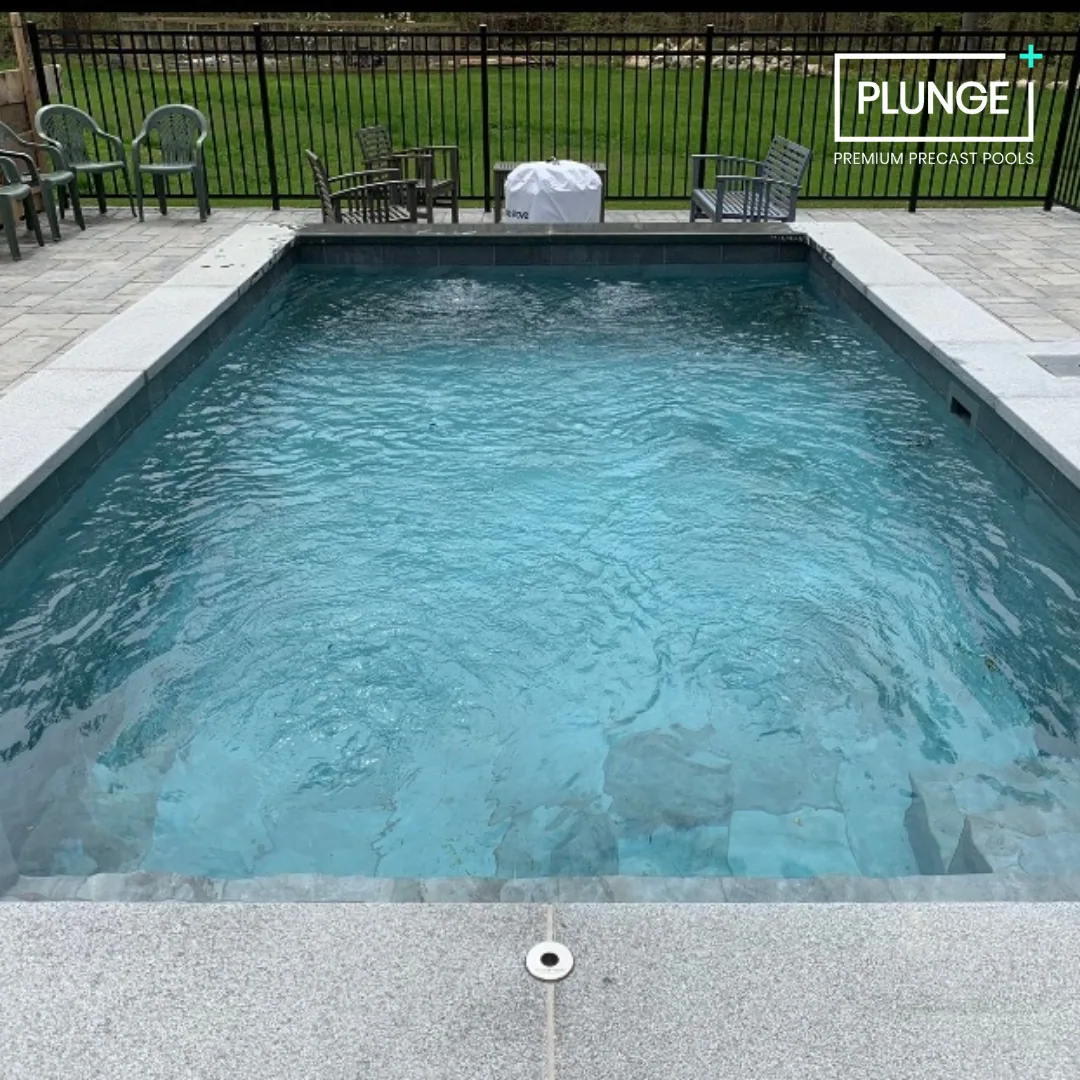 A rectangular private pool with clear blue water surrounded by a stone deck, with a black fence and green lawn in the background, and several chairs and benches on the deck. There is a cover over an object near the fence, and the image includes a log