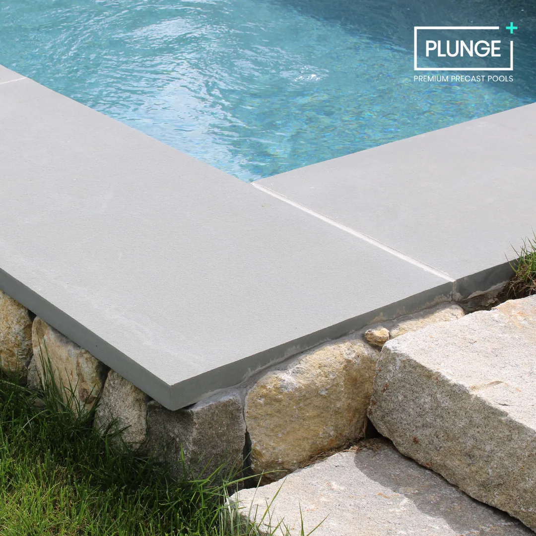 Close-up of a modern swimming pool with a concrete deck and stone border, showing clear water and a logo for 'PLUNGE' Premium Precast Pools.