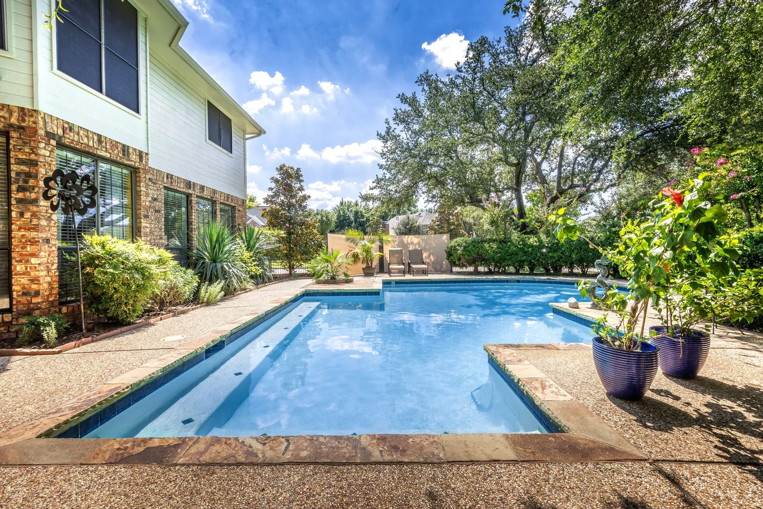 A backyard swimming pool with clear blue water, surrounded by plants and trees, with a house on the left and patio chairs at the far end, under a partly cloudy sky.