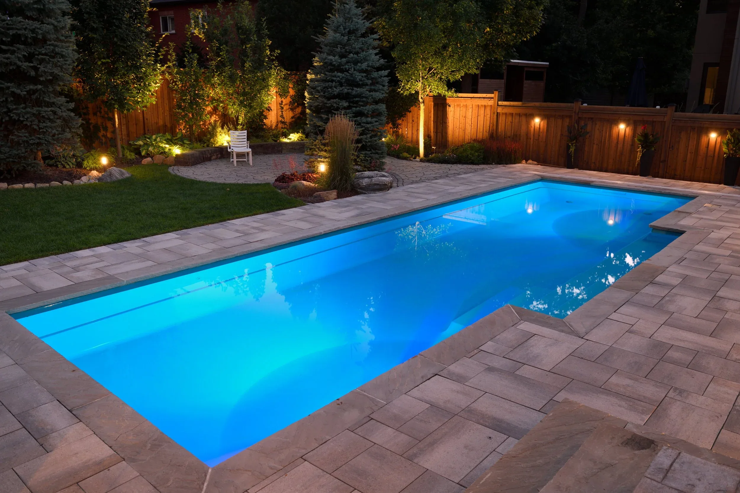 Nighttime view of a backyard with a lit blue swimming pool, surrounded by a stone patio, a grassy area, trees, and a wooden fence with outdoor lighting.