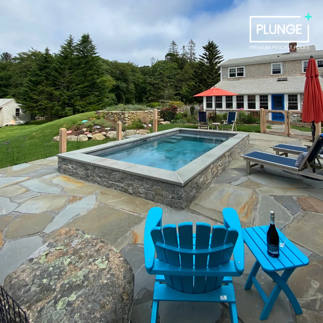 A backyard with a small rectangular pool, surrounded by a stone patio, with two blue Adirondack chairs, a matching side table with a bottle of champagne, and an orange umbrella near the house. There are trees and a garden in the background.