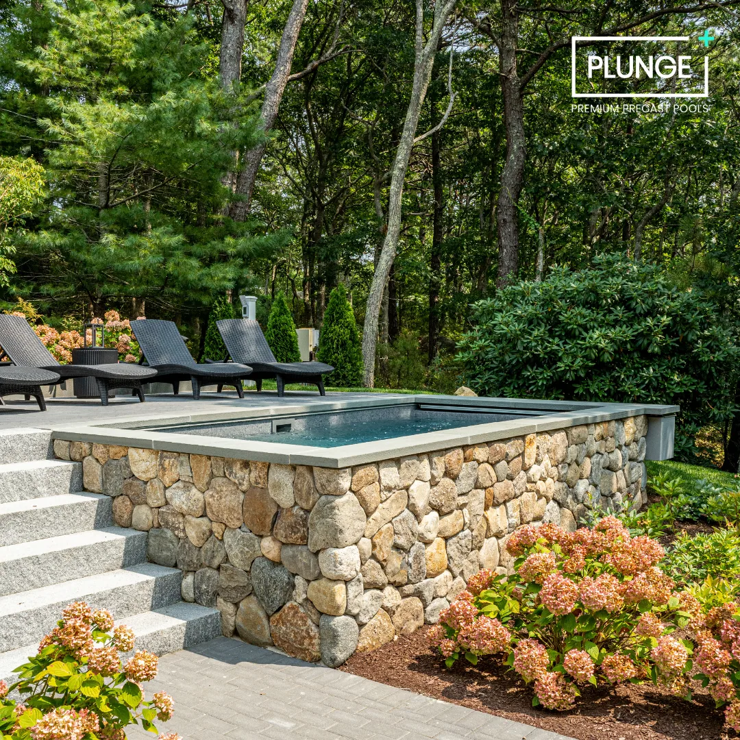 A backyard with a hot tub on a stone platform, surrounded by lush greenery and colorful flowers, with four black lounge chairs and trees in the background.