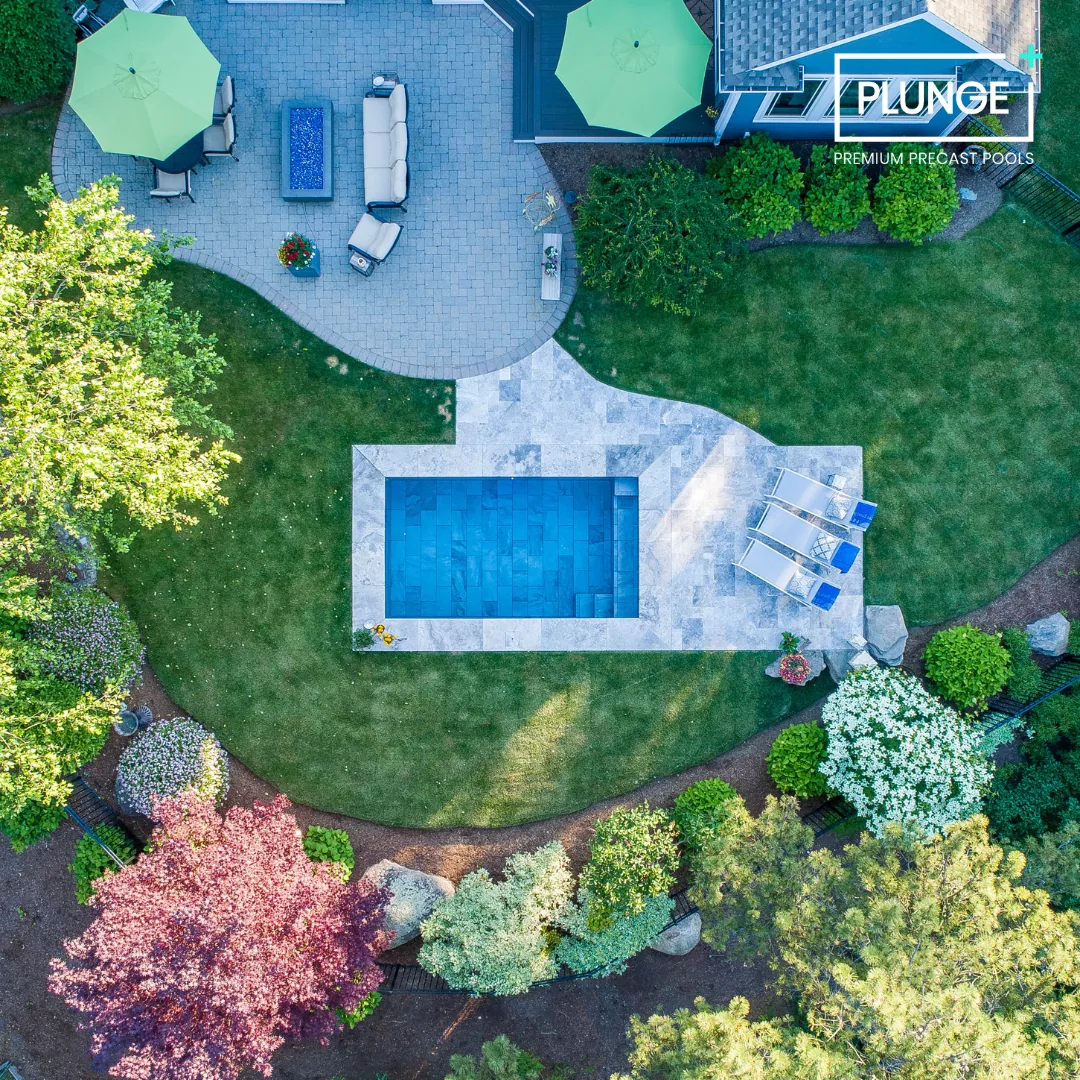 An aerial view of a backyard with a small pool, surrounding stone patio with lounge chairs and umbrellas, a sitting area, and lush greenery including trees and flowering bushes.