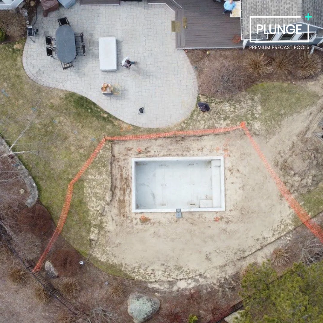 An aerial view of a backyard with a patio and an under-construction swimming pool, surrounded by grass and trees, with workers present.