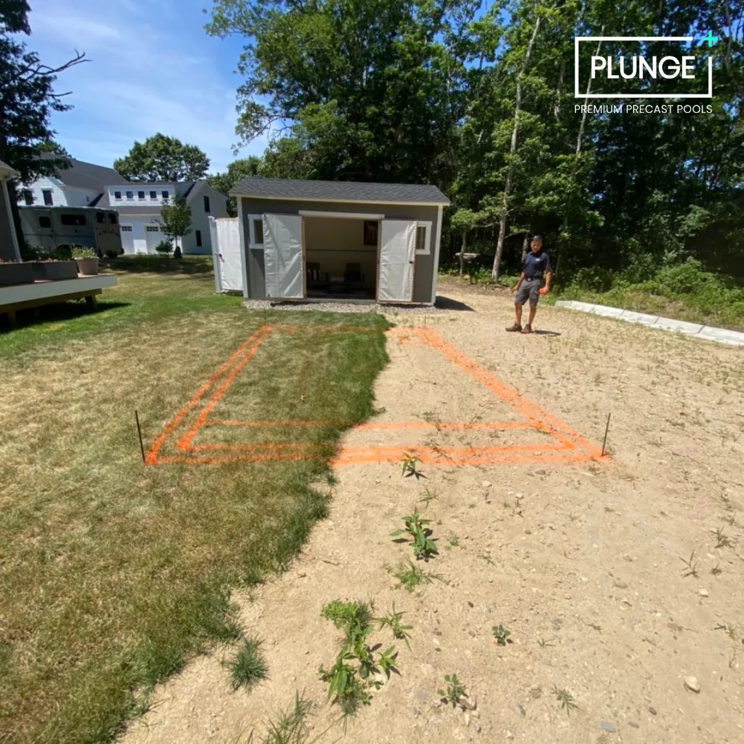 A backyard with a shed and a man walking on a dirt and grass area with orange spray paint markings for construction or landscaping work. There are trees and houses in the background, and a logo with the text 'PLUNGE Premium Precast Pools' in the top 