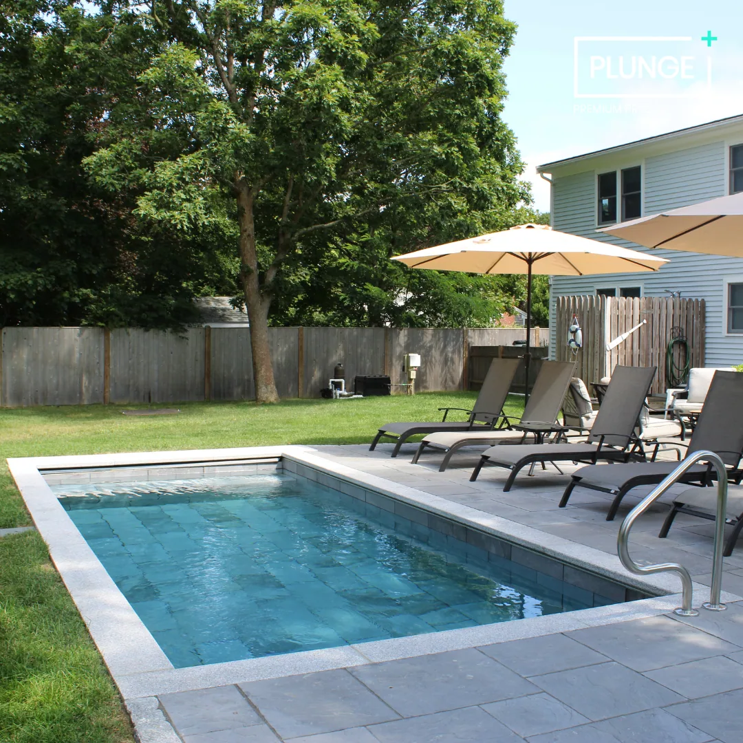 A backyard swimming pool with lounge chairs and patio umbrellas, surrounded by a grassy yard and a wooden fence, with trees in the background.