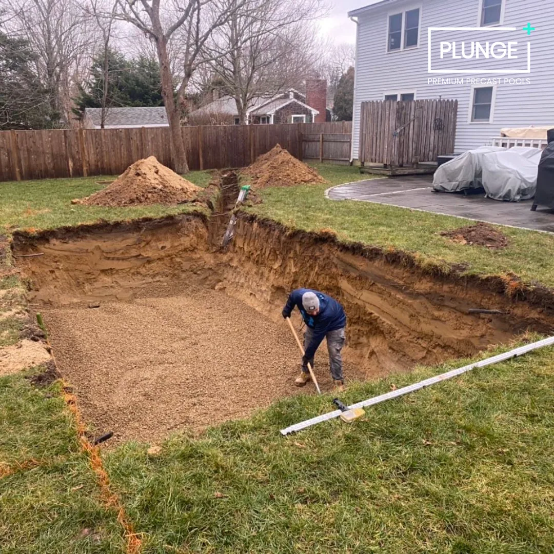 A person working in a backyard trench, installing a pool with gravel or sand. Piles of dirt, a hose, and construction tools are nearby. A fenced yard with a house, trees, and outdoor furniture covered with a tarp are in the background.