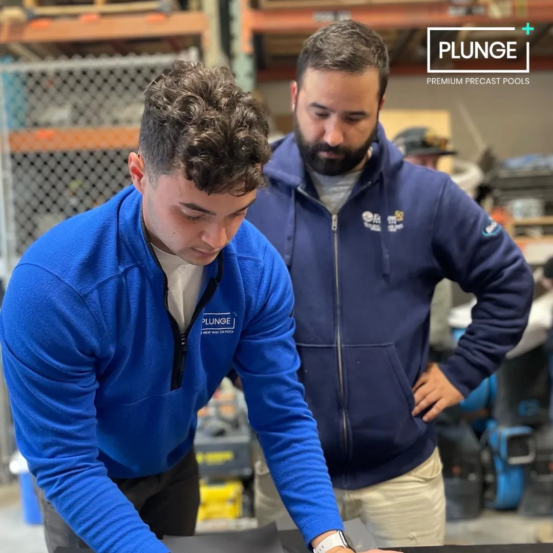 Two men in blue jackets working together in a workshop, with shelves and tools in the background, at a place called Plunge, premium precast pools.