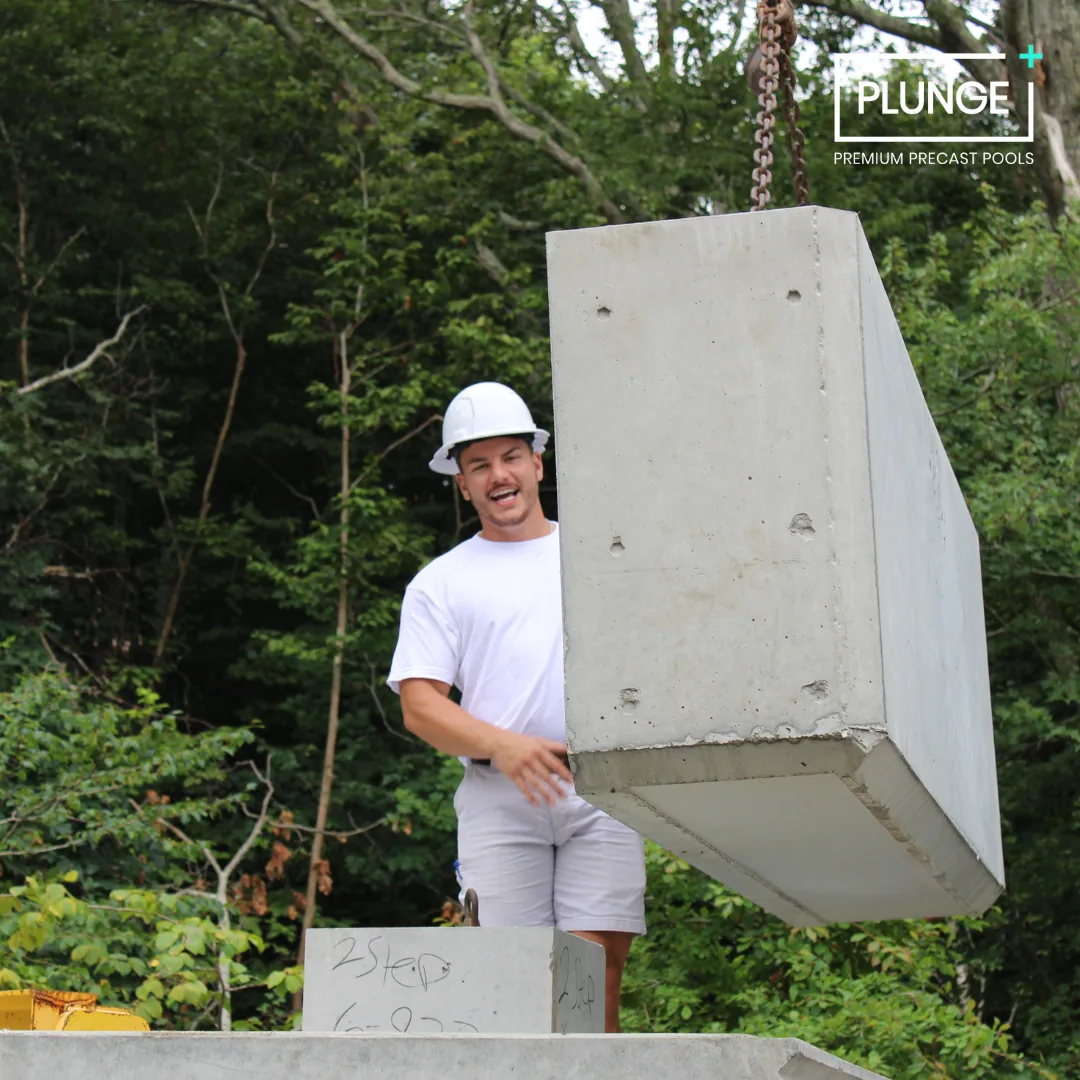 A construction worker in a white helmet and white shirt standing next to a large precast concrete pool wall being lifted by a crane outdoors surrounded by trees.
