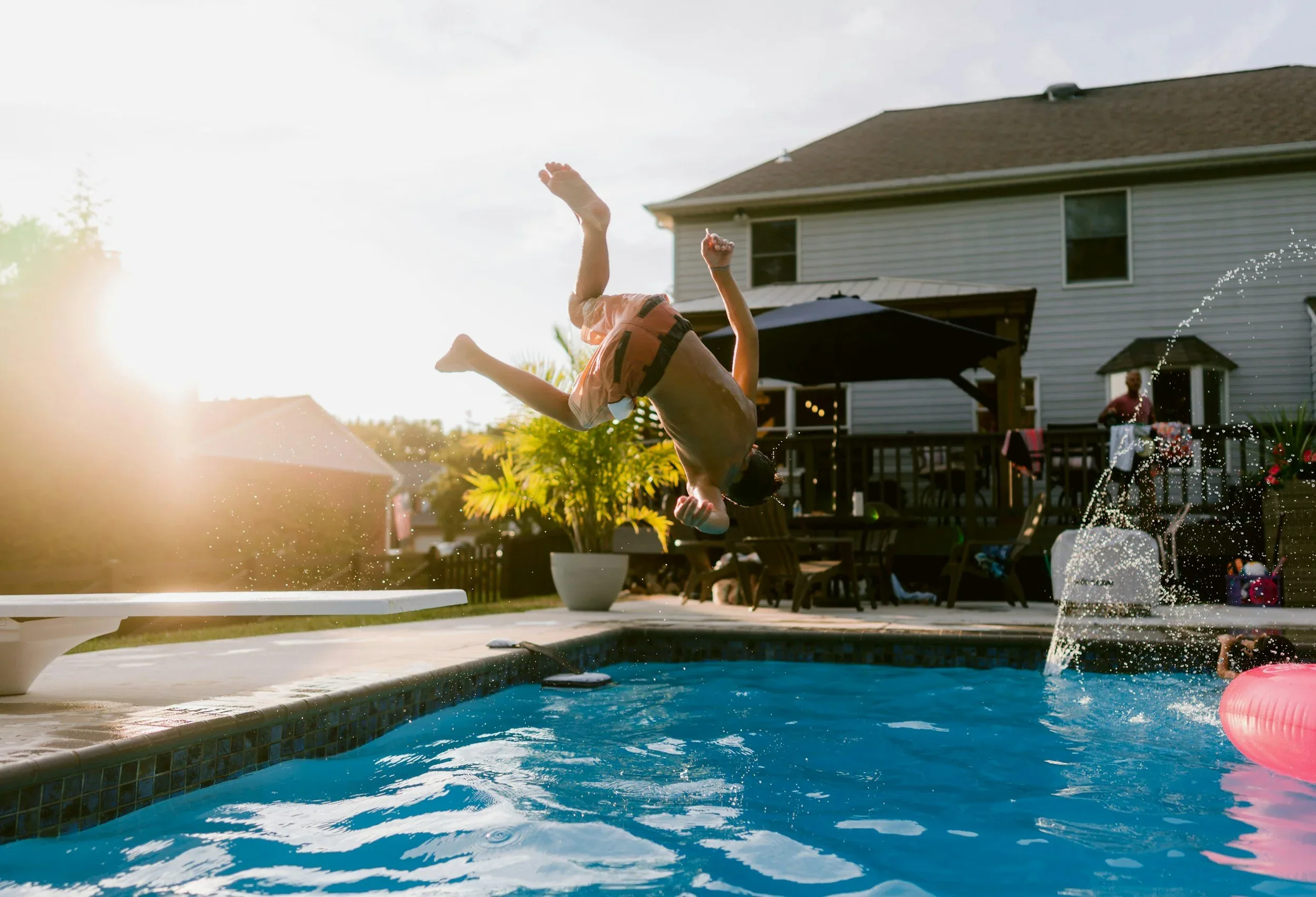 Child flipping into a backyard swimming pool during sunset, with a house and porch in the background, water splashing around.