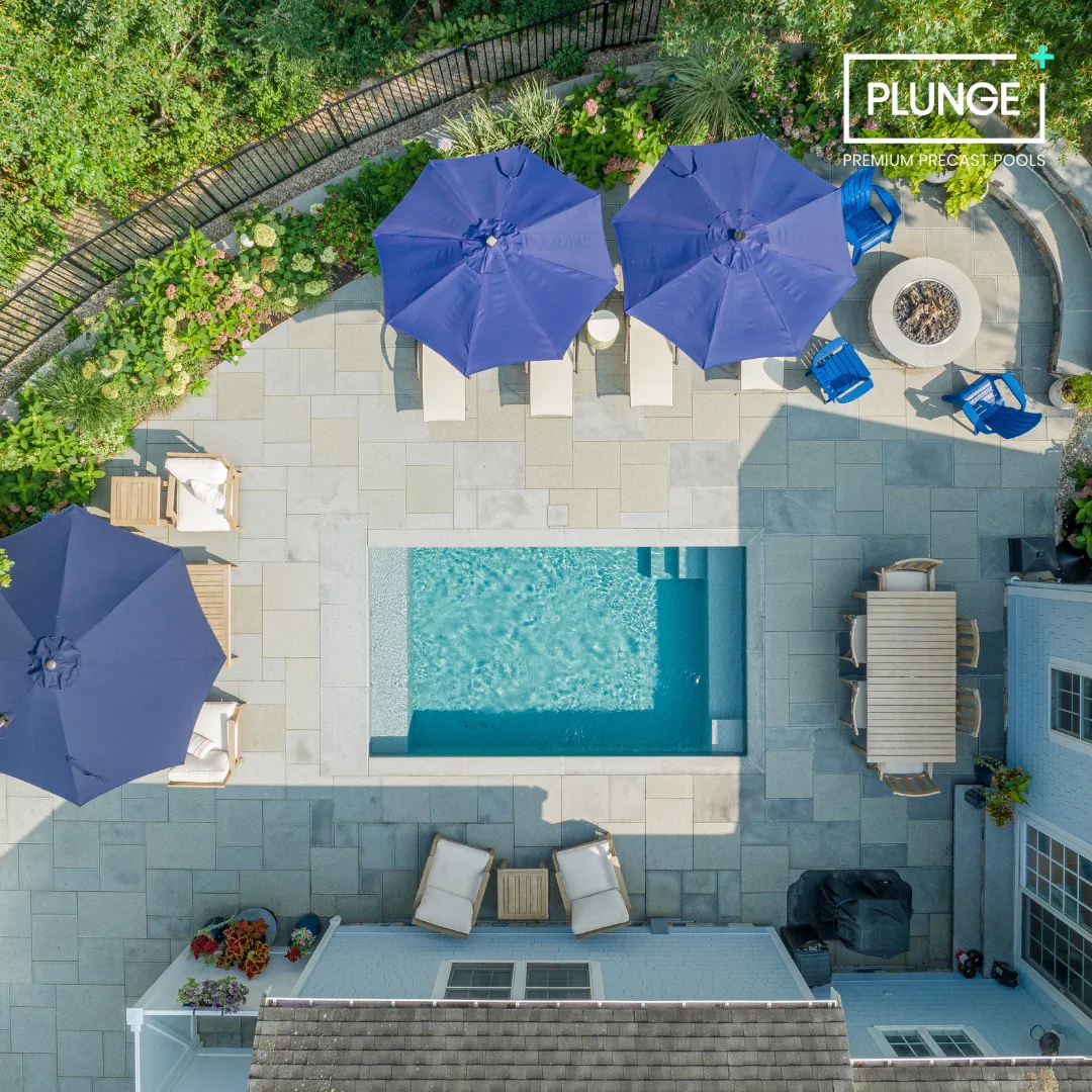 Aerial view of a backyard with a small swimming pool, surrounded by patio furniture, blue umbrellas, and a fire pit, with garden plants along the fence.