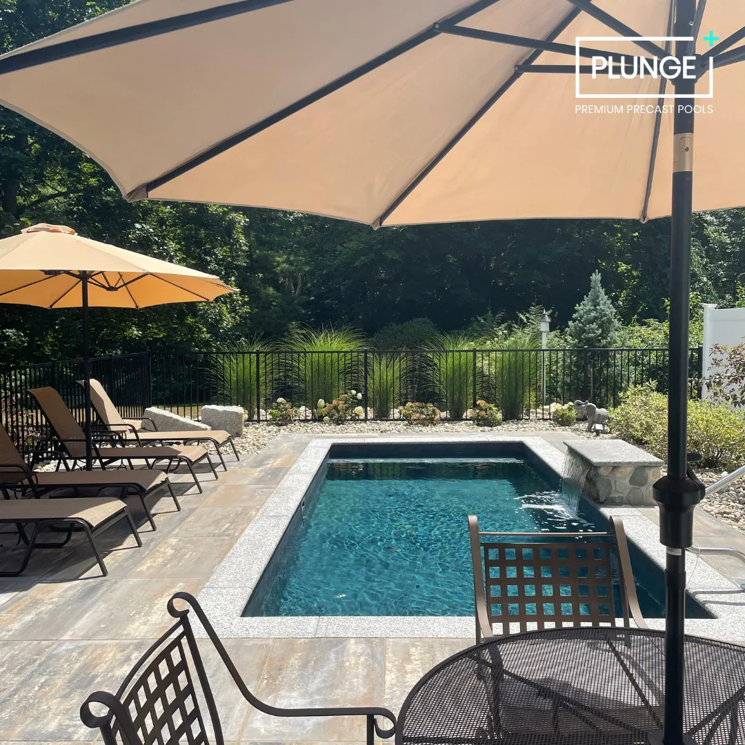 A backyard with a rectangular swimming pool, surrounded by lounge chairs, large umbrellas for shade, and lush greenery with trees and shrubs in the background.