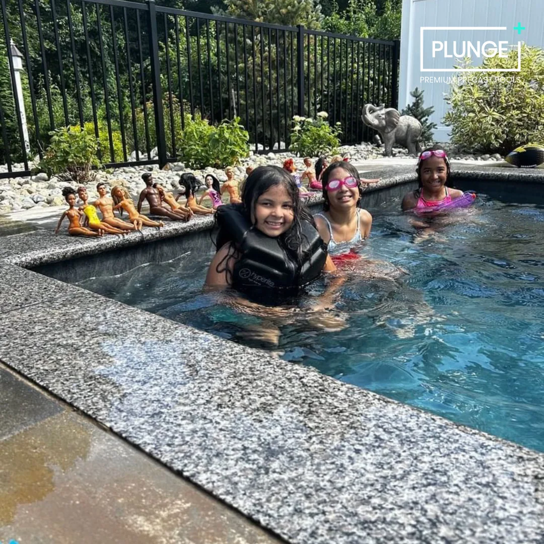 Three young girls are in a backyard swimming pool, smiling and enjoying the water. The girl on the left is wearing a life vest, while the other two are in swimwear with goggles. On the pool's edge, there are small dolls and toys arranged in a line. A small elephant sculpture and some plants are visible in the background."